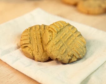 May include: Two round, golden-brown peanut butter cookies with a distinct crisscross fork pattern pressed into the top surface. The cookies are resting on a folded white fabric napkin, with additional blurred cookies visible in the background.