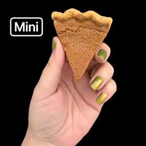 May include: A mini, triangular-shaped pastry with a light brown color and a wavy crust. The word "Mini" is displayed in a white box with a black background. The pastry is held against a black background.