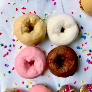 May include: Four frosted donuts in different colors: beige, white, pink, and brown. The donuts are arranged in a circle on a white surface with colorful sprinkles.