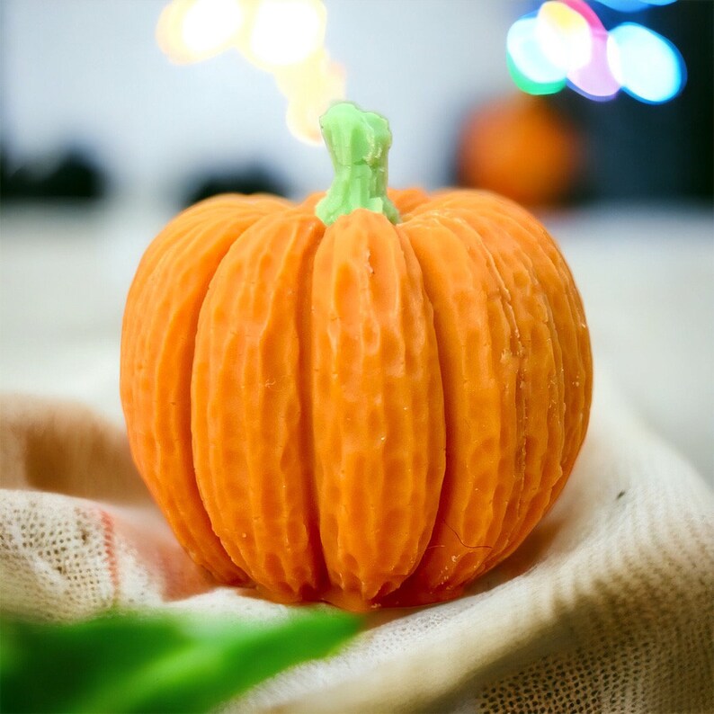 May include: A small, orange pumpkin with a green stem. The pumpkin has a textured surface and is sitting on a white and brown fabric.