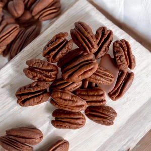 Könnte beinhalten: Nahaufnahme eines Stapels Pecan-Hälften auf einer weißen Holzoberfläche. Die Pecanüsse sind hellbraun und haben eine leicht glänzende Oberfläche.