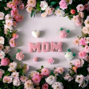 May include: A display of pink and white flowers encircles the word "MOM" in pink lettering. Decorative rose and heart-shaped embellishments are placed around the text, forming a celebratory and festive arrangement.