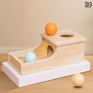 May include: A light-colored wooden ball drop toy featuring a rectangular base with a ramp and a top platform containing a circular hole. The set includes three smooth spheres in orange, light blue, and beige, with the DiasByDia logo visible.