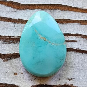 May include: A polished, teardrop-shaped turquoise gemstone with a glossy finish. The stone has a light blue hue with subtle brown veining. The gemstone is set against a rustic, white and brown wooden surface.