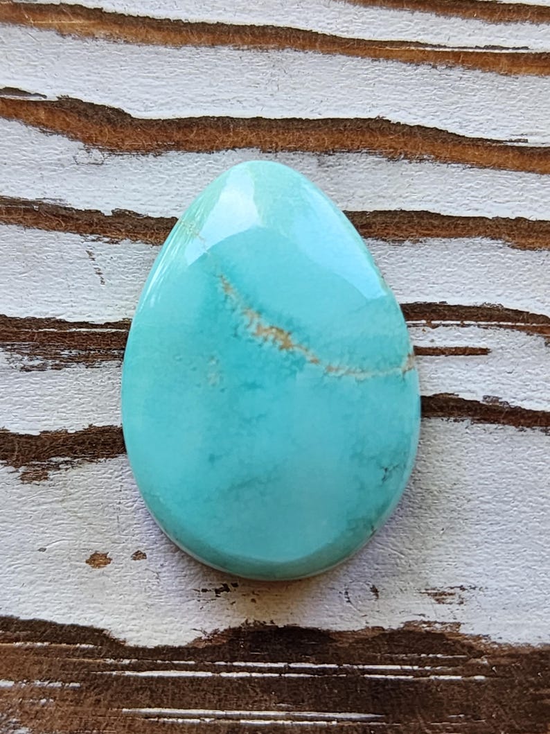 May include: A polished, teardrop-shaped turquoise gemstone with a glossy finish. The stone has a light blue hue with subtle brown veining. The gemstone is set against a rustic, white and brown wooden surface.