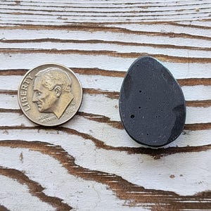 May include: A dark grey, teardrop-shaped stone with small, light-coloured inclusions. A U.S. dime coin is next to the stone for size comparison. The stone is on a white-painted, distressed wood surface.