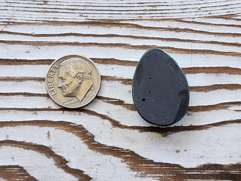May include: A dark grey, teardrop-shaped stone with small, light-coloured inclusions. A U.S. dime coin is next to the stone for size comparison. The stone is on a white-painted, distressed wood surface.