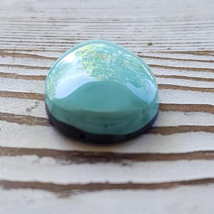 May include: A smooth, teardrop-shaped teal cabochon with a black base. The polished surface reflects the surrounding environment. The cabochon is set against a light brown, wooden background. This item is suitable for jewellery making.