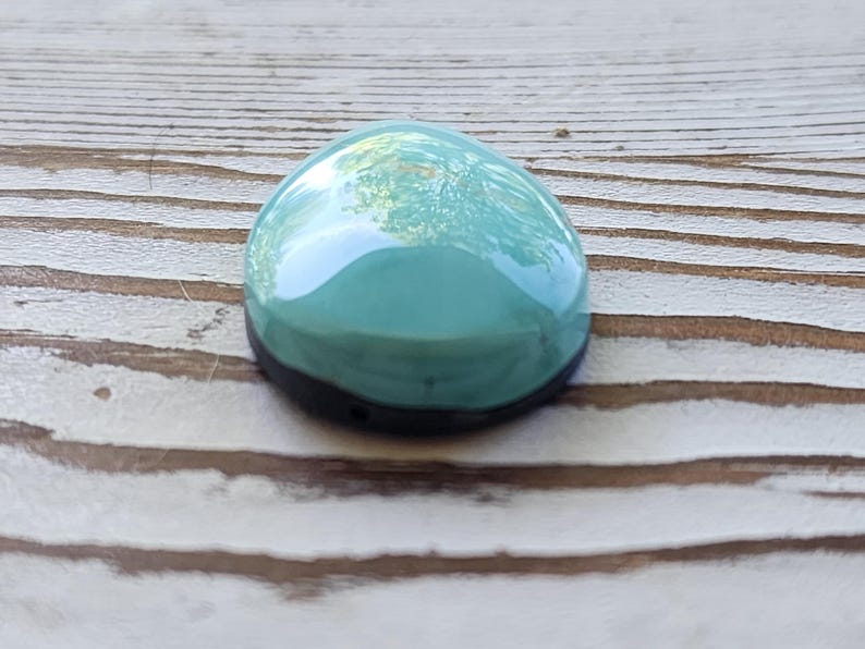 May include: A smooth, teardrop-shaped teal cabochon with a black base. The polished surface reflects the surrounding environment. The cabochon is set against a light brown, wooden background. This item is suitable for jewellery making.