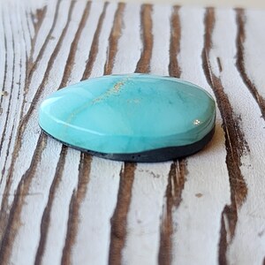 May include: A polished, oval turquoise gemstone with a dark grey base. The stone has a smooth, glossy surface with subtle gold inclusions. The gemstone is set against a weathered, white-painted wooden surface.
