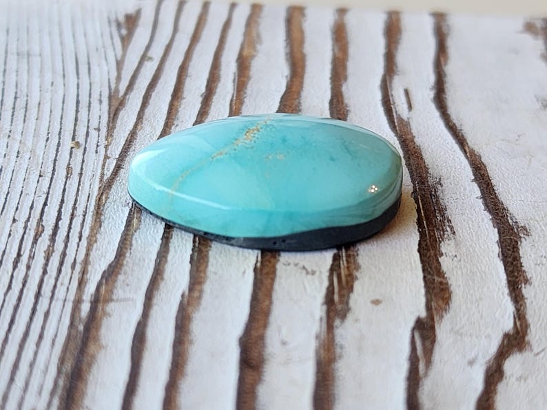 May include: A polished, oval turquoise gemstone with a dark grey base. The stone has a smooth, glossy surface with subtle gold inclusions. The gemstone is set against a weathered, white-painted wooden surface.