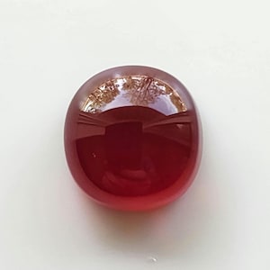 May include: A polished, oval-shaped gemstone with a deep red hue. The cabochon cut stone reflects light, creating a glossy surface. The gemstone is set against a white background.