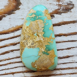 May include: A polished, teardrop-shaped gemstone with a turquoise base color and intricate gold and tan veining patterns. The stone has a smooth, glossy surface and is set against a rustic, weathered wood background.