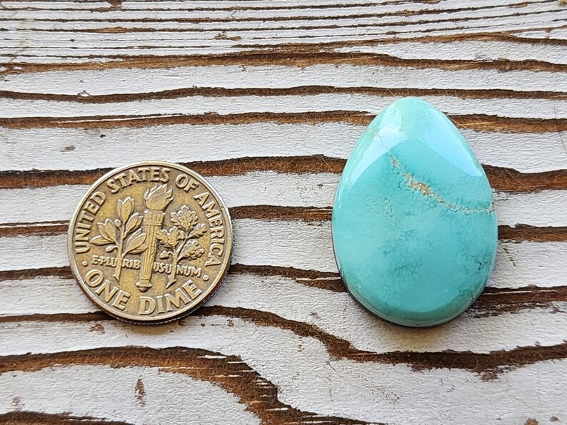 May include: A polished, teardrop-shaped turquoise gemstone with natural veining, next to a United States one dime coin. The coin is approximately 1.8 cm in diameter. The gemstone is a vibrant blue-green colour.