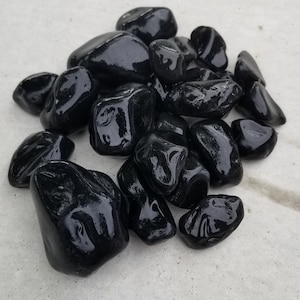 May include: A pile of smooth, polished black river rocks. The stones have a glossy, reflective surface and vary in size and shape. They are arranged on a light gray concrete surface.