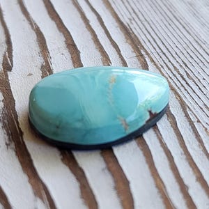 May include: A polished, oval turquoise gemstone with a dark rim. The stone has a smooth, glossy surface with a marbled appearance, displaying shades of blue and hints of brown. The gemstone is set against a weathered, light-coloured wooden surface.