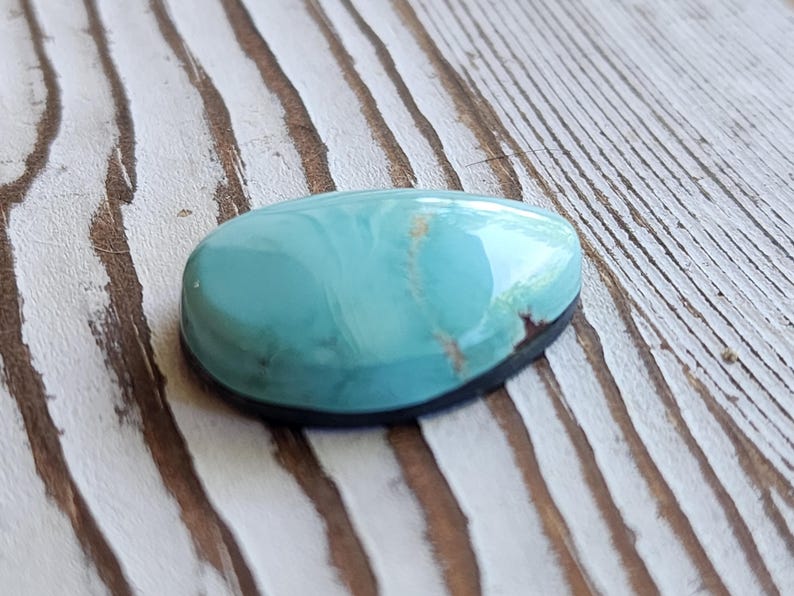 May include: A polished, oval turquoise gemstone with a dark rim. The stone has a smooth, glossy surface with a marbled appearance, displaying shades of blue and hints of brown. The gemstone is set against a weathered, light-coloured wooden surface.