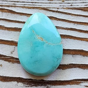 May include: A polished, teardrop-shaped turquoise gemstone. The stone has a smooth, glossy surface with a light brown vein running through the centre. The gemstone is set against a weathered, light-coloured wooden surface.