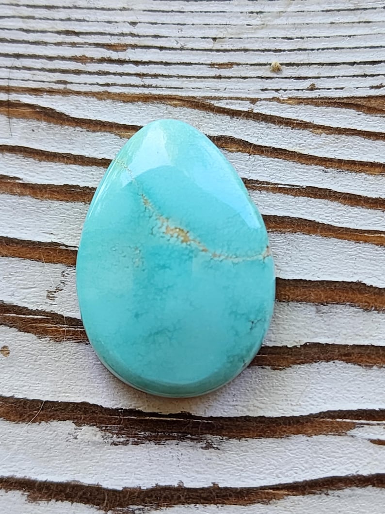 May include: A polished, teardrop-shaped turquoise gemstone. The stone has a smooth, glossy surface with a light brown vein running through the centre. The gemstone is set against a weathered, light-coloured wooden surface.