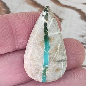 Puede incluir: Una gema en forma de lágrima con una franja central azul turquesa. La piedra tiene una base de color crema con una red de finas vetas blancas. La parte superior de la piedra tiene un patrón verde oscuro y negro.