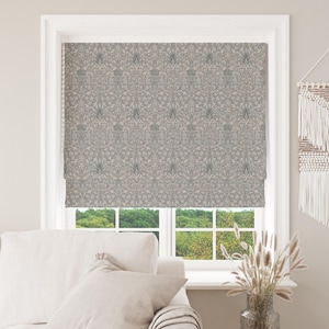 May include: A Roman blind with a floral design in grey and beige tones is fitted within a white window frame. The window offers a view of a green landscape. A white sofa and wall hanging are also visible.