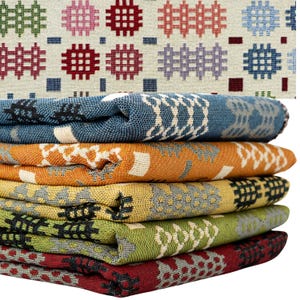 May include: A stack of five folded woven fabrics featuring a repeating geometric grid pattern. The top fabric is cream with multicoloured grid motifs, followed by layers in blue, orange, yellow, green, and red, each with textured patterns.
