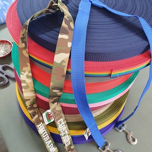 Könnte beinhalten: Ein Stapel bunter Hundeleinen mit verschiedenen Mustern und Farben, darunter Tarnmuster, Regenbogen und einfarbige Farben. Die Leinen haben Metallclips und sind mit den Namen "Rhabarber" und "Cieland" beschriftet.