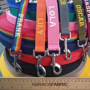Puede incluir: Una pila de correas para perros de colores con nombres personalizados, incluyendo "Frank", "Lily", "Lola", "Cooper", "Oscar" y "Jasper". Las correas están hechas de nylon tejido y tienen clips de metal plateado. Una regla de madera con el texto "ABRACAFABRIC" está en primer plano.