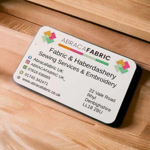 May include: A white business card with black text and a colorful logo. The text reads "ABRACAFABRIC Fabric & Haberdashery Sewing Services & Embroidery" and includes contact information: Abracafabric UK, ABRACAFABRIC UK_, 07818 636665, 01745 342471, www.abracafabric.co.uk. The address is 22 Vale Road, Rhyl, Denbighshire, LL18 2BU.