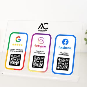 以下が含まれることがあります： 透明なアクリル製の看板に3枚のカードが置かれています。カードにはQRコードとGoogle、Instagram、Facebookのソーシャルメディアロゴが掲載されています。カードのテキストには、「Googleでレビューを残してください」、「Instagramでフォローしてください」、「Facebookでフォローしてください」と記載されています。