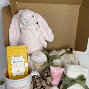 Pode incluir: Uma caixa de presente cheia de um coelhinho de pelúcia rosa, uma caneca com a palavra "mama", chá, uma bomba de banho, um tubo de loção, uma scrunchie de cabelo, uma toalha pequena e uma colher dourada. A caixa é marrom e os itens estão dentro.
