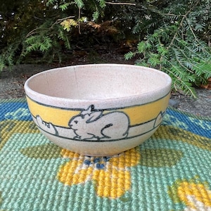 May include: A ceramic bowl with a cream-colored interior and a yellow band featuring a black outline of a rabbit. The bowl sits on a colorful, patterned rug, with green foliage in the background. The bowl is a decorative item.