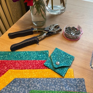 Può includere: Un set di cinque pochette in tessuto colorate con un motivo a cerchi. Le pochette sono disposte in pila, con la pochette verde in basso e la pochette turchese in alto. Una piccola pochette turchese con una chiusura a scatto si trova al centro della pila. Nell'immagine sono presenti anche un paio di pinze per punzonare i fori in metallo e un contenitore di bottoni a pressione in metallo.
