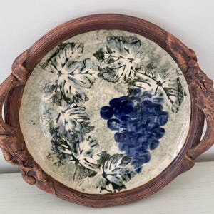 May include: A round ceramic serving tray with a brown rim and handles. The tray features a central design of a cluster of dark blue grapes and surrounding green and white grape leaves. The rim and handles have a textured, wood-like appearance.