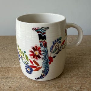 May include: A white ceramic mug featuring a large, stylized letter J decorated with intricate, multi-colored floral patterns in shades of red, blue, green, and orange. The mug has a smooth, cylindrical body with a handle on the right side.