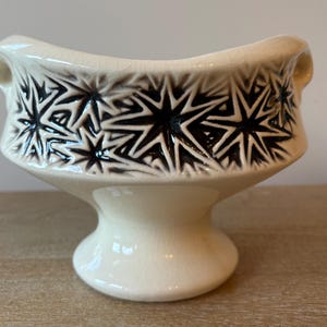 May include: Cream-colored ceramic bowl with a pedestal base and two small handles. The bowl's exterior features a repeating pattern of dark starburst designs. The bowl has a glossy finish and sits on a wooden surface.