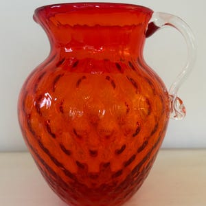 May include: A vibrant orange glass pitcher with a clear glass handle. The pitcher has a textured surface with a pattern of small, raised dots. The pitcher is a decorative item.