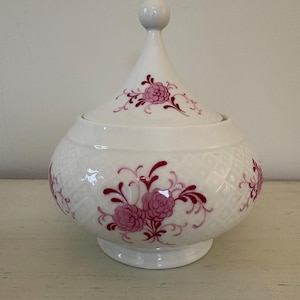 May include: A white ceramic lidded jar with a bulbous body and a conical lid. The jar is decorated with a pink floral pattern and a diamond-shaped texture. The lid has a small, round knob.