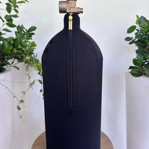 May include: A black neoprene tank cover with a gold zipper, designed to fit a gas cylinder. The top features a brass valve and a gray knob. The cylinder is placed on a small, round wooden table, with greenery in the background.