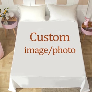 Pode incluir: Um lençol de baixo branco com o texto "Custom image/photo" em castanho avermelhado. Duas almofadas brancas são visíveis no topo da cama. A cama está num quarto bem iluminado.