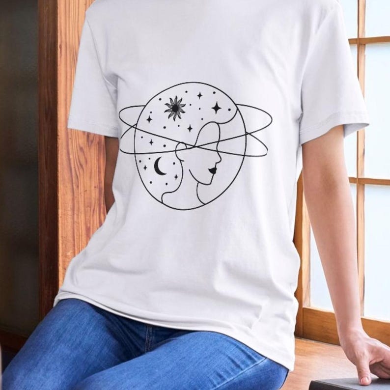 May include: White t-shirt with a black line drawing of a woman's face inside a circle with stars, a crescent moon, and a sun with rays.