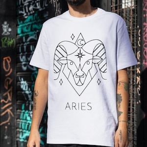 May include: White t-shirt with a black line drawing of an Aries zodiac symbol, including a ram's head, crescent moon, and stars. The word "ARIES" is printed below the symbol.