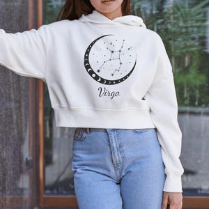 May include: White cropped hoodie with a black graphic of the Virgo zodiac sign and the word "Virgo" printed below. The hoodie has a drawstring hood.