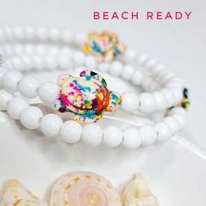 Puede incluir: Pulseras de cuentas blancas con abalorios de tortuga coloridos. Los abalorios presentan un salpicado de colores brillantes. El texto "BEACH READY" se muestra en letras rosas en la parte superior de la imagen. Se ven conchas marinas en la parte inferior.