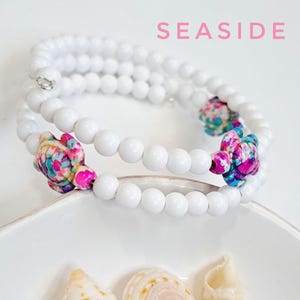 Può includere: Braccialetto di perline bianche con accenti di tartarughe colorate. Il braccialetto presenta un design a spirale con perline bianche e due ciondoli a forma di tartaruga con motivi rosa, blu e verdi. La parola "SEASIDE" è in rosa.