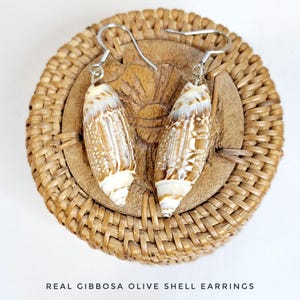 May include: A pair of earrings made from real olive shells with silver hooks. The shells are a light brown color with white stripes. The earrings are displayed on a woven basket.