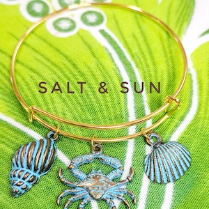 May include: Gold bangle bracelet with three charms: a blue seashell, a blue crab, and a blue seashell. The bracelet has the text "SALT & SUN" on it.