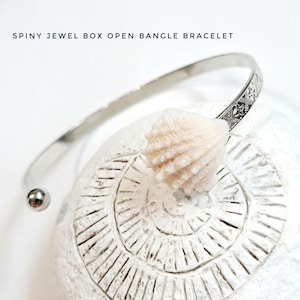 May include: A silver open bangle bracelet with a decorative star pattern on one end. The bracelet rests on a white, textured surface with a seashell. The text at the top reads: SPINY JEWEL BOX OPEN BANGLE BRACELET.