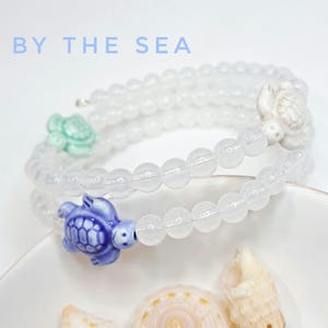 Könnte beinhalten: Ein weißes Perlenarmband mit drei Schildkrötenanhängern in Blau, Grün und Weiß. Das Armband ist gewickelt und liegt auf einer weißen Oberfläche. Der Text "BY THE SEA" steht oben in Hellblau.