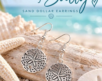 May include: Silver-toned sand dollar earrings featuring a detailed five-petal star pattern and textured surface. The earrings hang from silver-toned hooks. The text "stay Salty" and "SAND DOLLAR EARRINGS" is displayed above the jewelry.